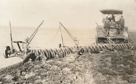 A sepia photo of a farm machine plowng a dirt field.